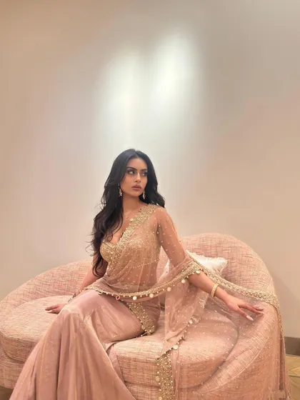 Seated on a curved sofa, this shot showcases the flowing sharara pants and the intricately embroidered cape-style dupatta.