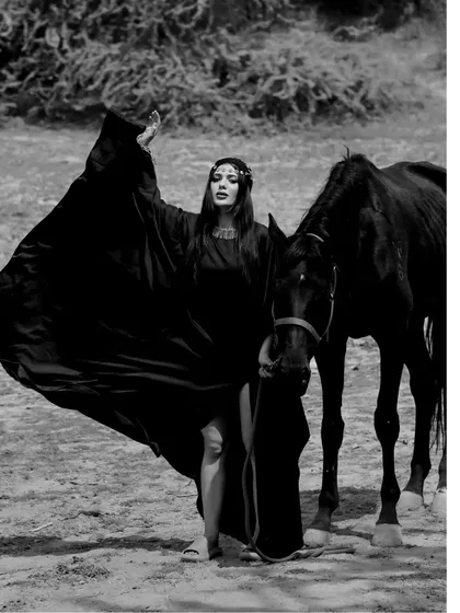 A full-length black and white shot of a model with a flowing cape standing next to a black horse, creating a majestic and powerful image.