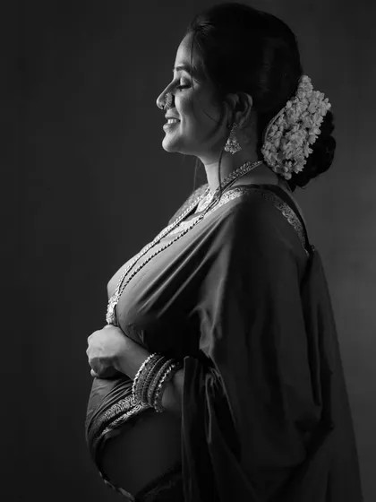 An intimate black and white side-profile portrait, capturing the joyful smile of a mother-to-be with her hair adorned with flowers.