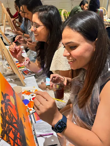 Smiles, sips, and painting. A candid shot of participants enjoying a relaxed afternoon, adding the final touches to their sunset paintings with a drink in hand.