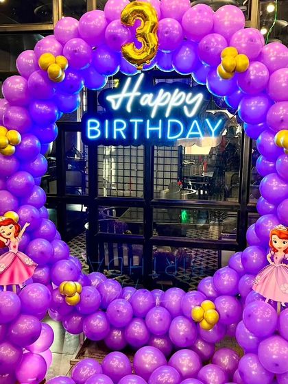 A magical princess-themed party for a 3rd birthday. This setup includes a full ring of purple balloons, a "Happy Birthday" neon sign, and cutouts of Princess Sofia.