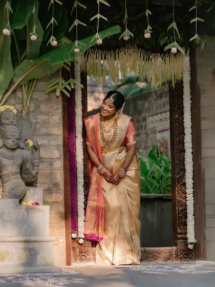 A bride, in a golden saree, peeks out from a decorated doorway, her happy smile lighting up the frame.