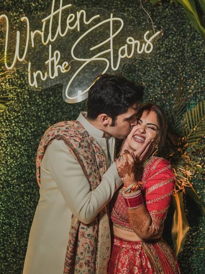 A playful and adorable portrait of the groom kissing his laughing bride on the cheek, in front of a 'Written in the Stars' neon sign.