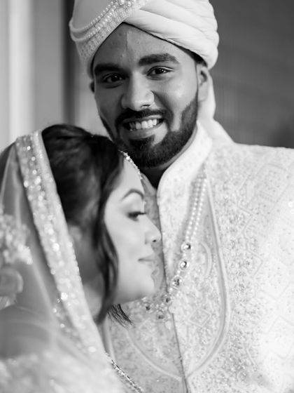 A candid black and white portrait where the groom is smiling at the camera while his bride rests her head on his shoulder.