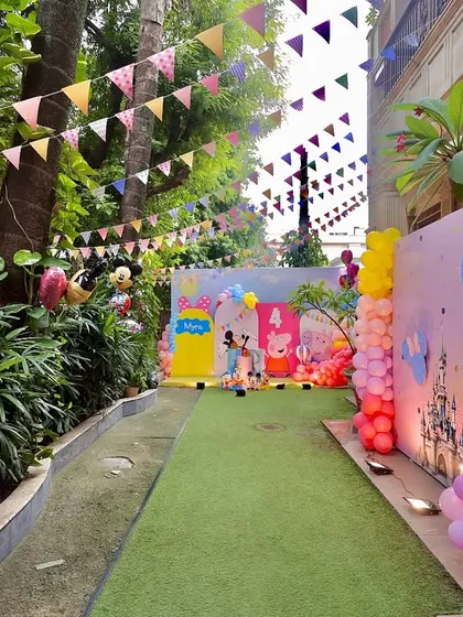 A view of the Peppaland party entrance pathway, lined with lush greenery, festive bunting, and colorful balloon arrangements, creating an exciting journey for arriving guests.