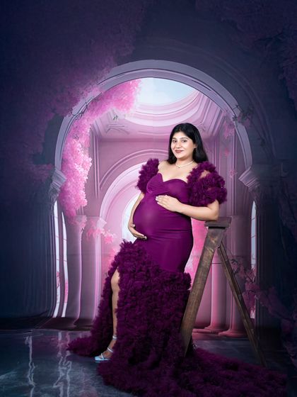 A regal maternity portrait of a woman in a deep purple gown, seated on a ladder in a dramatic, floral-arched studio.