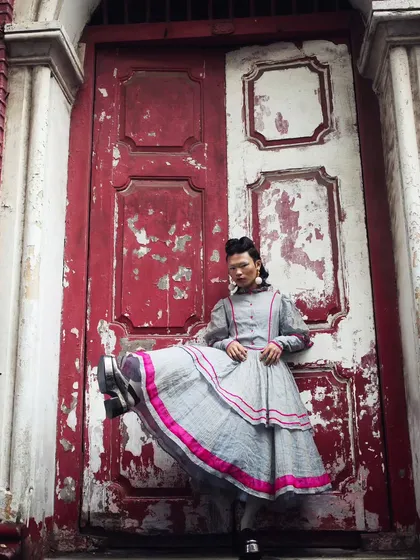 A full-length shot from the "RIOT" series, showcasing the entire look against the dramatic backdrop of a weathered red and white door. The pose is both elegant and defiant.