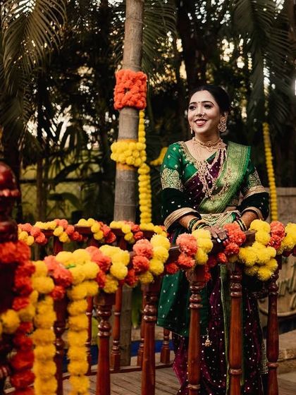 A Maharashtrian bride stands behind a railing adorned with marigold garlands. Her elegant green saree and joyful expression perfectly capture the festive spirit of the wedding day.