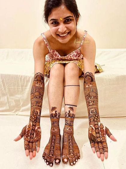 A happy bride showing off her complete bridal mehendi, with intricate designs on both her hands and feet.
