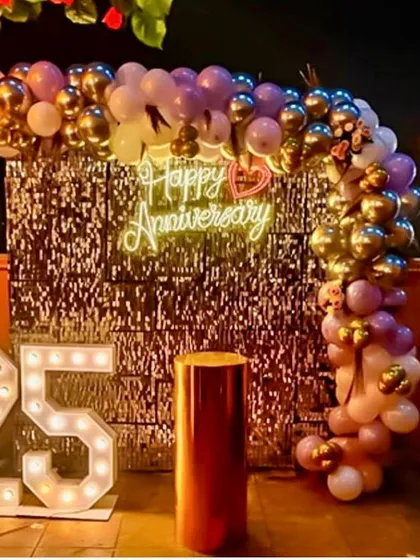 An outdoor terrace setup for a 25th anniversary. This design features a silver sequin wall that sparkles at night, a balloon arch in purple and gold, and large '25' marquee numbers.
