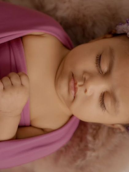 A close-up of a sleeping newborn wearing a purple floral headband, wrapped in a matching purple cloth.