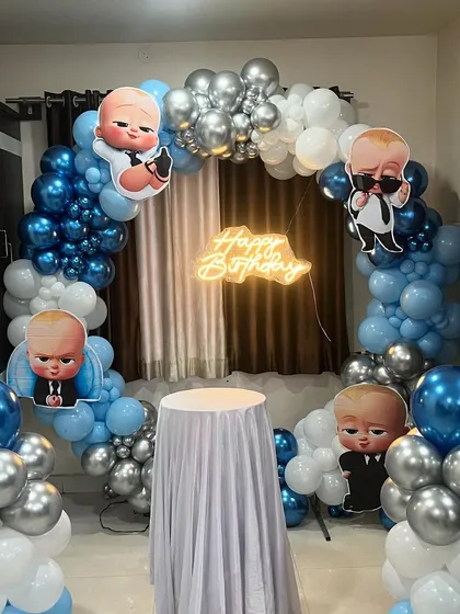 A simple yet effective Boss Baby theme for a home party. The setup includes a balloon arch backdrop, character cutouts, and a 'Happy Birthday' neon sign.