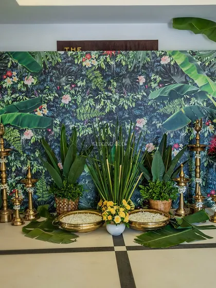 A stunning Haldi backdrop blending nature and heritage. A tropical-print wall is accented with lush banana leaves, traditional brass lamps, and bowls of yellow flowers.