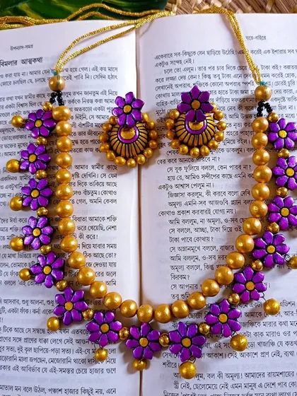 This floral necklace features purple flowers on a string of golden beads, artfully arranged on a book of Bengali poetry. It comes with beautiful matching earrings.