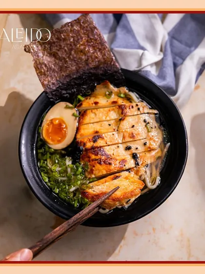 A close-up of our comforting chicken ramen bowl. Notice the perfectly seared chicken, the rich jammy egg, fresh greens, and a sheet of nori, all swimming in a deeply flavorful broth.