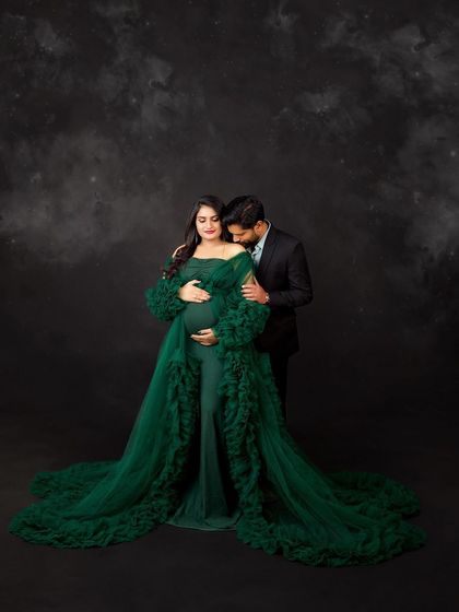 A dramatic and intimate maternity portrait. The deep green gown and the partner's gentle kiss create a sense of romance and anticipation against a dark, textured backdrop.