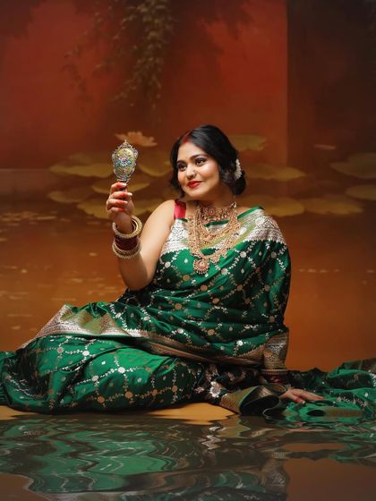 A reflective moment captured with a traditional 'aarshi' mirror. This pose, set against a painted lotus pond backdrop, evokes a sense of timeless Indian artistry.