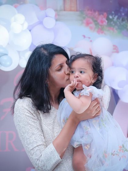 A mother's gentle kiss for her daughter amidst a sea of pastel balloons. A sweet, quiet moment during a busy party.