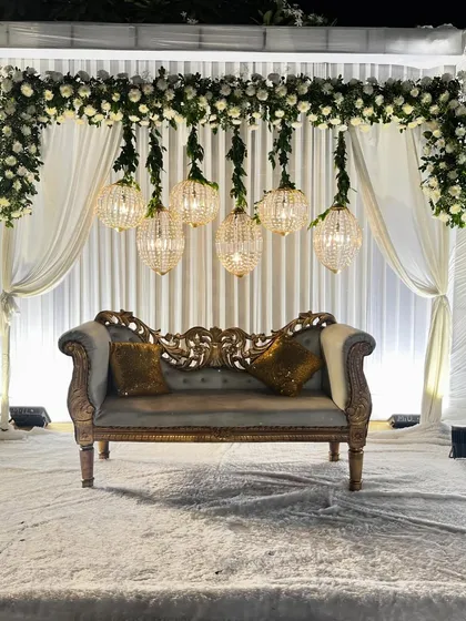 A classic and elegant Walima stage setup. The decor features hanging crystal chandeliers and a vintage-style sofa, framed by white drapes and florals.