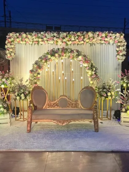 An outdoor evening engagement setup with a beautiful floral arch and candle-like lighting. The vintage sofa and white carpet create a romantic focal point.