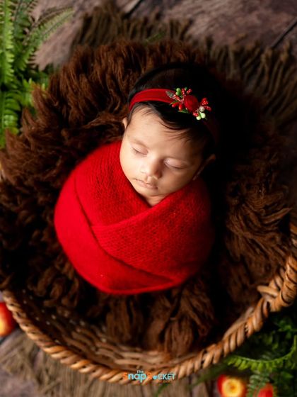 This baby girl is wrapped in a festive red swaddle, nestled in a rustic basket with greenery. This setup has a warm, almost holiday-like feel.