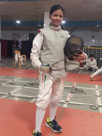 A young fencer takes a moment to pose after a training session, holding her mask and sabre. She is geared up and ready for the challenges of the sport.
