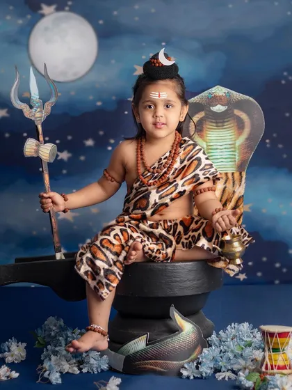 A little girl dressed as Lord Shiva, holding a trishul and bell, looks directly at the camera with a powerful gaze.