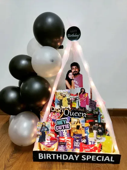 This 'Birthday Special' canopy hamper creates a grand statement. The tent-like structure is adorned with balloons and fairy lights, enclosing a curated selection of treats and a personalized photo cutout.