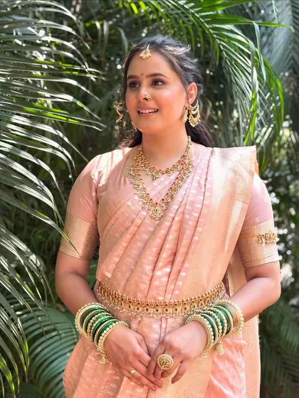 A lovely three-quarter shot showing the bride against a backdrop of green palms. The natural light beautifully highlights the dewy finish of the makeup and the elegance of her traditional attire.