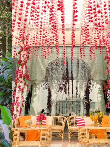 Hanging strings of red and pink flowers create a beautiful curtain of color for this intimate Haldi seating area. The bamboo furniture and vibrant cushions add to the fresh and festive atmosphere.