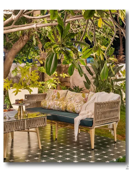 An outdoor lounge at night, with the woven sofa and glass table illuminated by the warm ambient light. The surrounding tropical foliage creates a cozy and intimate atmosphere.