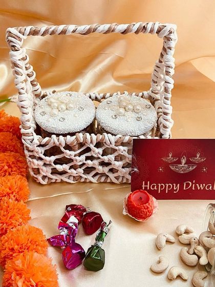 A unique woven basket holding two decorative jars, perfect for gifting homemade sweets or dry fruits. This hamper is accompanied by chocolates and a diya candle.