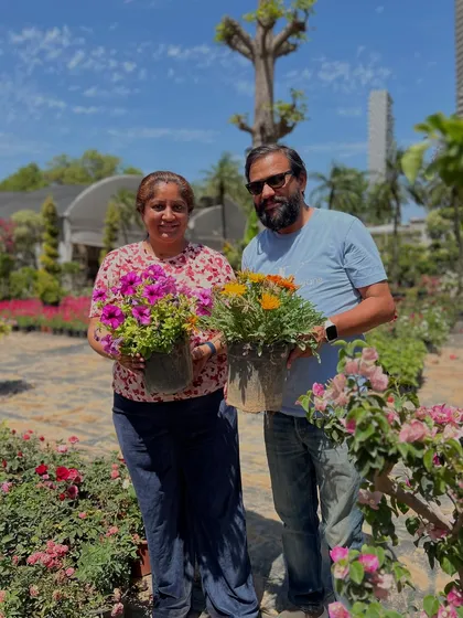 A happy customer couple holding their new flowering plants, surrounded by the beautiful blooms at our nursery.