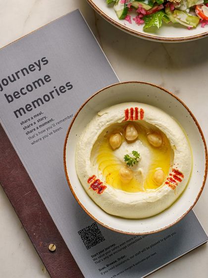 A bowl of creamy hummus, elegantly styled with whole chickpeas and a drizzle of olive oil. The restaurant's menu in the background adds context, making this a great shot for promotional use.