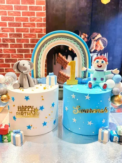 A set of two separate first birthday cakes for twins, connected by a beautiful pastel rainbow.