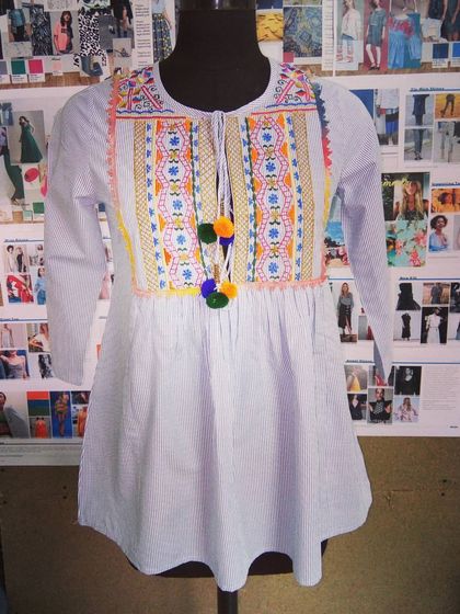 A fun and trendy boho-chic top on a mannequin. This striped top features colorful tribal-inspired embroidery and playful pompom tassels, perfect for a casual yet stylish look.
