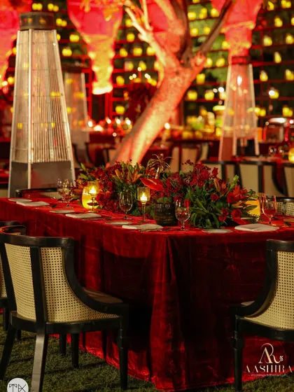 A closer view of the dining table, showing the rich red velvet tablecloth, cane-back chairs, and the beautiful floral centerpiece under the warm light of a patio heater.