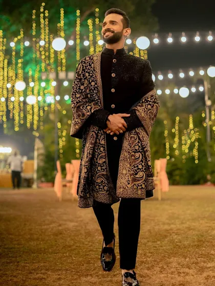 A stylish portrait of the groom in a classic black sherwani with an intricately designed shawl, walking confidently at his outdoor reception.