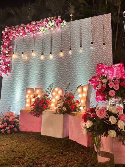 A romantic photo backdrop for an outdoor event. The design features a wall with a geometric pattern, hanging Edison bulbs, and the couple's names in large, illuminated marquee letters.