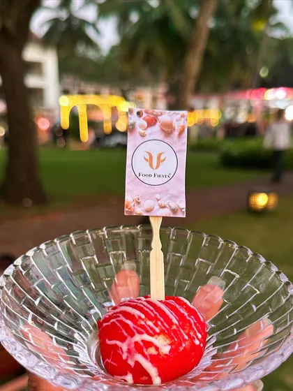 A single, vibrant red dessert ball drizzled with white chocolate, held up against the backdrop of an outdoor event in Goa. It's a perfect, bite-sized piece of happiness.