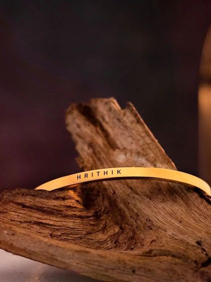 A gold-plated cuff engraved with 'HRITHIK', resting on a piece of driftwood. The warm, rustic setting complements the rich gold tone of the bracelet.