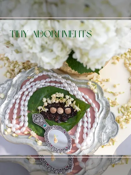 Tiny adornments for a welcome ceremony, featuring a betel leaf with supari, a pearl necklace, and a vintage hand mirror on a silver tray.