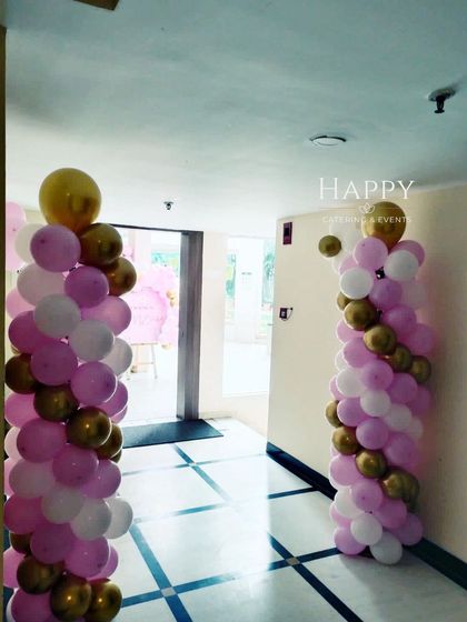 Elegant balloon pillars in pink, white, and gold lining the hallway. These decorative elements guide guests to the main event area while enhancing the party's ambiance.