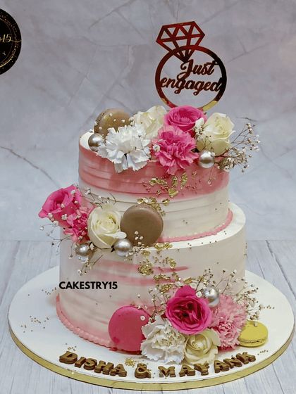 A stunning two-tier choco-butterscotch engagement cake. This 5kg creation features a blush pink watercolor effect, fresh flowers, macarons, and a "Just Engaged" topper.