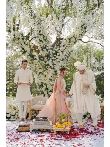 A sacred moment during an intimate garden ceremony. The couple stands before a stunning tree of life, its branches laden with white flowers, as they prepare to take their seven steps together, surrounded by a carpet of red rose petals.