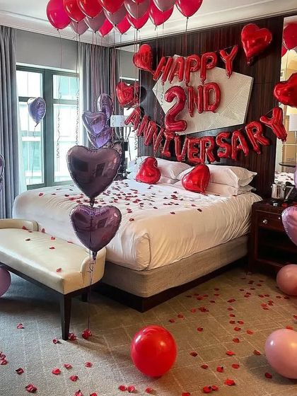 A luxurious 2nd-anniversary hotel room decoration with red and purple heart-shaped foil balloons, rose petals, and ceiling balloons.