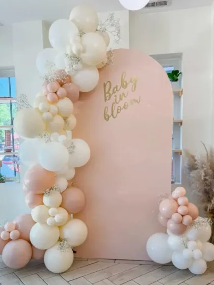 A "Baby in Bloom" shower theme with a soft pink arched backdrop. The elegant balloon garland in shades of blush and cream, with tiny baby's breath flowers, creates a delicate and beautiful look.