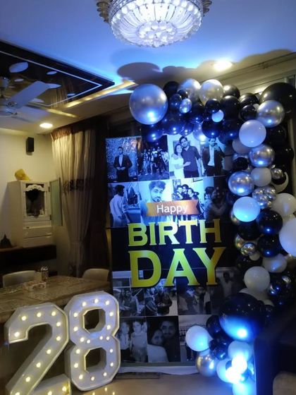 A stylish photo collage backdrop for a 28th birthday celebration at home. The black and white photos are paired with marquee numbers and a black and silver balloon garland.