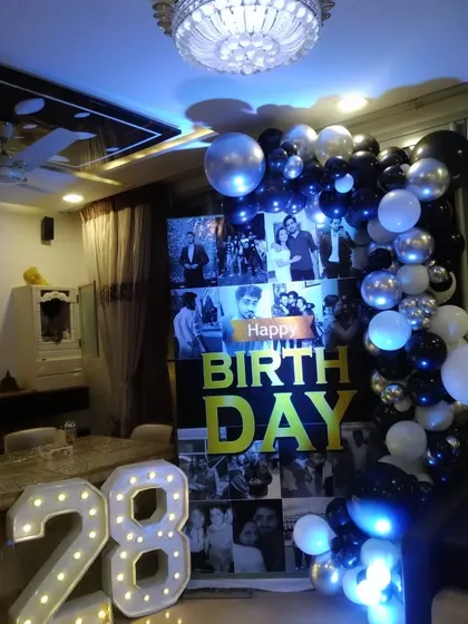 A stylish photo collage backdrop for a 28th birthday celebration at home. The black and white photos are paired with marquee numbers and a black and silver balloon garland.