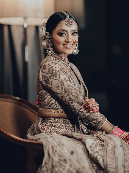 A classic, elegant bridal portrait. The bride is seated, smiling warmly at the camera, showcasing her intricate jewelry and hairstyle.